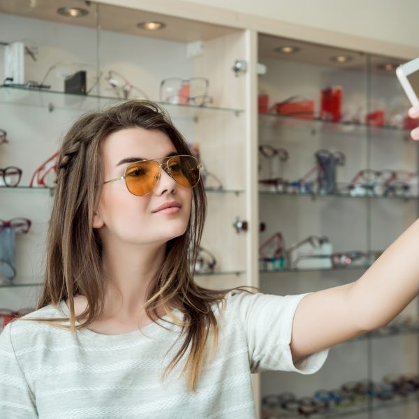 Attractive woman went on shopping alone, making selfie while trying on new stylish sunglasses in optician shop, sending photo to friend so she will say her opinion. Girl bought trendy eyewear. Copy space